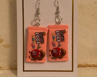 May include: A pair of pink M&M's Eggs earrings. The earrings are made of polymer clay and feature a miniature M&M's Eggs candy bar. The earrings are attached to silver hooks.