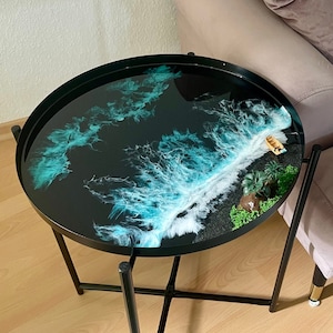 Könnte beinhalten: Ein runder schwarzer Beistelltisch mit einem schwarzen Metallgestell. Die Tischplatte zeigt eine Ozeanszene aus Harz mit blauen und weißen Wellen, schwarzem Sand und einer kleinen Palme.