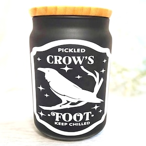 以下が含まれることがあります： 木製の蓋が付いた黒い瓶で、「PICKLED CROW'S FOOT KEEP CHILLED」というテキストとカラスのイラストが描かれた白いラベルが付いています。瓶は円筒形で、ガラス製のように見えます。