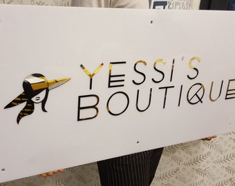 May include: A white sign with gold lettering that reads "Yessi's Boutique". A stylised image of a woman wearing a hat is to the left of the text.