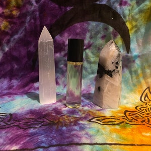 May include: Three items on a tie-dye fabric background: a clear glass roller bottle with a black cap, a white crystal point, and a white crystal point with black inclusions.
