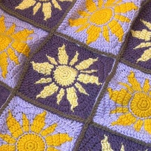 Peut inclure: Gros plan sur une couverture crochetée avec un motif de tournesol répété. Les tournesols sont jaunes, sur un fond violet, avec des bordures marron. La couverture faite à la main est un article décoratif et confortable.