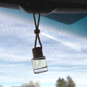 May include: A small, clear glass car air freshener bottle with a wooden cap and a hanging rope. The bottle is filled with a light-colored liquid and hangs from a car's rearview mirror against a blue sky.