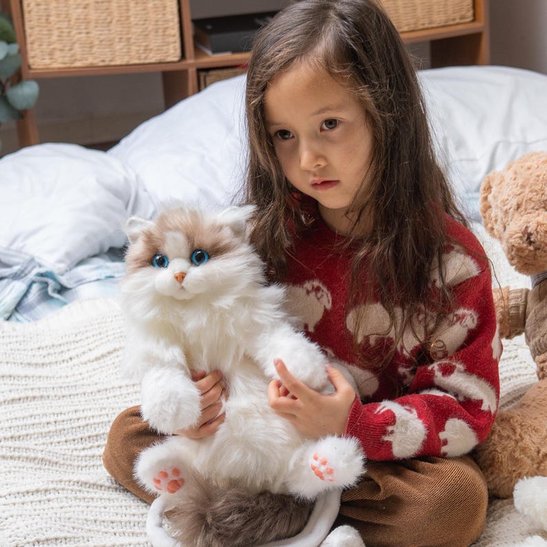 May include: A young girl in a red sweater with a white sheep pattern is sitting on a bed and holding a white fluffy cat plush toy. The cat has blue eyes and pink paw pads.