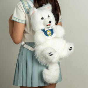 Puede incluir: Una mochila de oso de peluche blanco y esponjoso con un babero azul con un emblema dorado. El oso tiene huellas de patas negras y una lengua rosa. La mochila se lleva sobre el hombro.