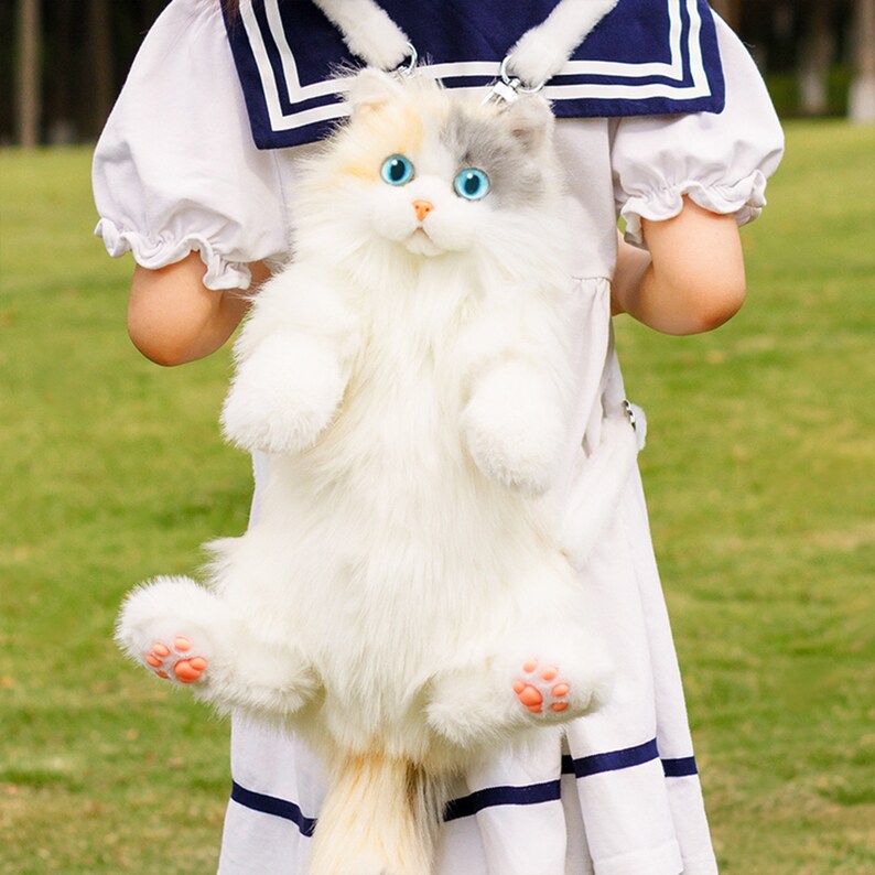 May include: A white and gray plush cat backpack with blue eyes. The cat is wearing a white and blue sailor collar.