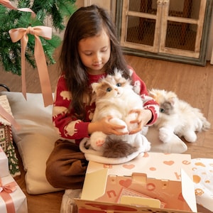 May include: A young girl in a red and white sweater holds a white and gray plush cat. The cat is sitting on a brown and white blanket. A second white and gray plush cat is lying on the blanket. The girl is sitting on a beige floor with a Christmas tree in the background.