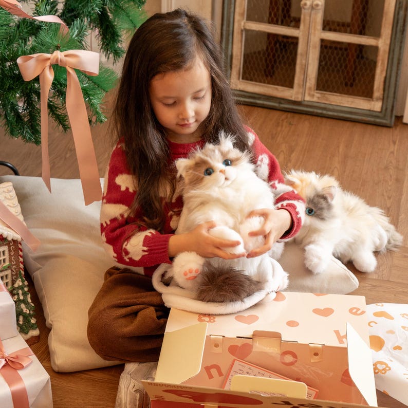 May include: A young girl in a red and white sweater holds a white and gray plush cat. The cat is sitting on a brown and white blanket. A second white and gray plush cat is lying on the blanket. The girl is sitting on a beige floor with a Christmas tree in the background.