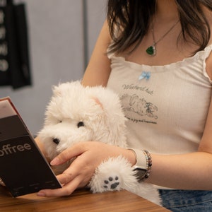 May include: A person is holding a white plush toy dog. The dog has a black nose and paw prints on its paws. The person is wearing a white tank top with the text "Weekends hub." and a blue bow. The person is also wearing a watch and a bracelet.