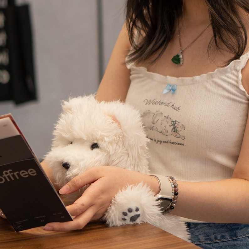 May include: A person is holding a white plush toy dog. The dog has a black nose and paw prints on its paws. The person is wearing a white tank top with the text "Weekends hub." and a blue bow. The person is also wearing a watch and a bracelet.