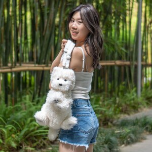May include: A white plush dog backpack with a white strap. A person is wearing the backpack and smiling at the camera.