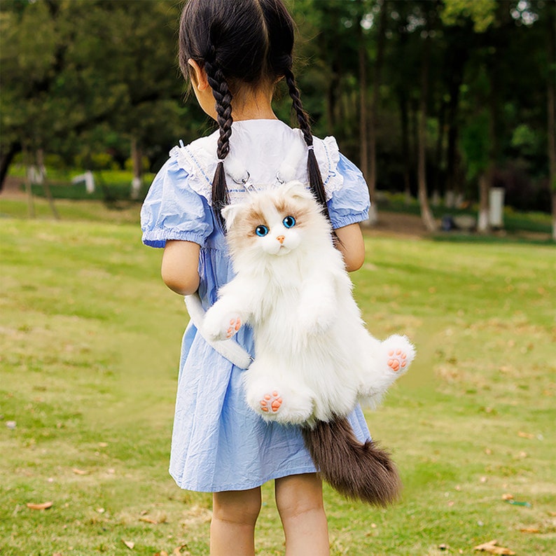 May include: A white and brown plush cat backpack with blue eyes. The cat is wearing a pink collar and has a long tail. The backpack is being worn by a child in a blue dress.