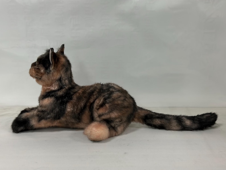 May include: A brown, black and white tabby cat plush toy is lying on its side with its head turned to the left. The cat has a long tail and is made of soft, plush fabric.