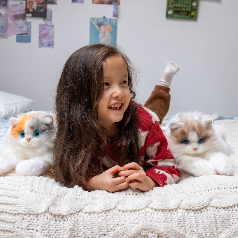 May include: A young girl with long brown hair is lying on a white blanket with two plush cats. One cat is white with blue eyes and the other is white, orange, and gray with blue eyes.