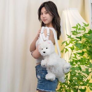 May include: A white plush dog backpack with a white strap. The backpack is being worn by a person with long dark hair.