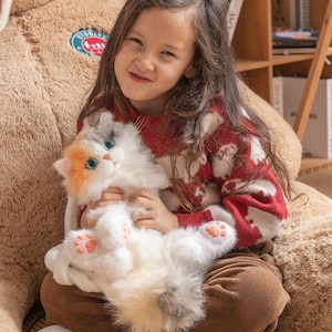 May include: A young girl wearing a red and white sweater holds a white, gray, and orange plush cat toy. The cat toy has blue eyes and pink paw pads.