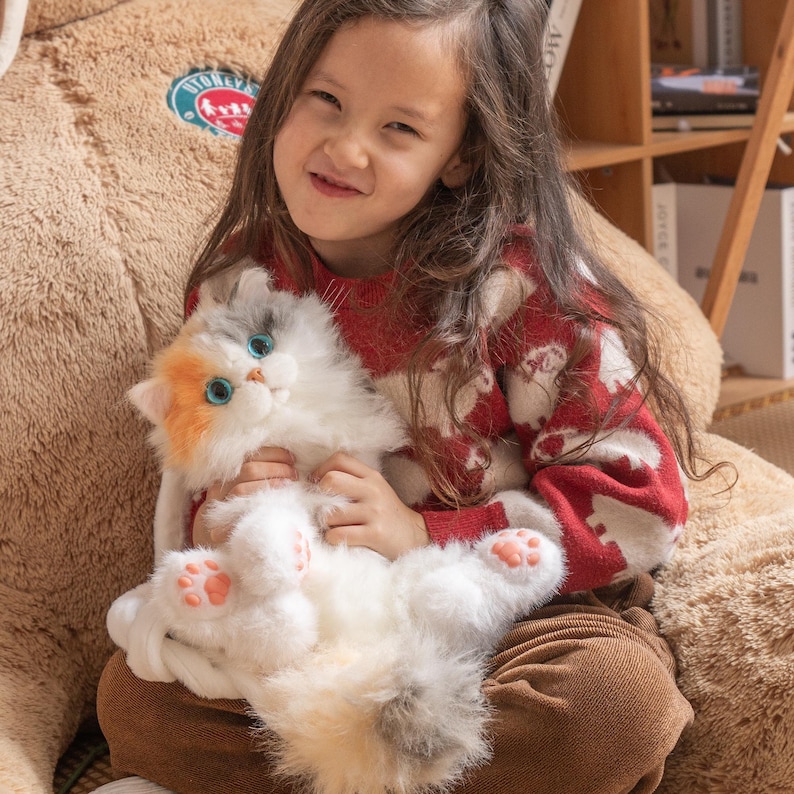 May include: A young girl wearing a red and white sweater holds a white, gray, and orange plush cat toy. The cat toy has blue eyes and pink paw pads.