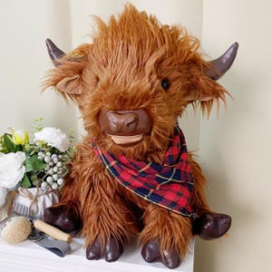 May include: A plush brown Highland cow toy with a red and black plaid bandana. The toy has a friendly face and is sitting on a white surface.