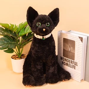 Può includere: Un gatto di peluche nero con occhi verdi e una collana di perle con un ciondolo di gemma verde. Il gatto è seduto su una superficie marrone chiaro accanto a un libro con il testo "Wonderful Homelife".
