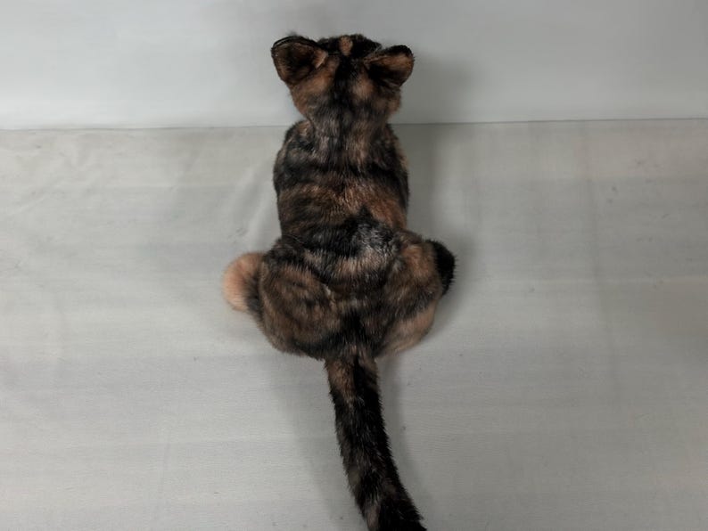 May include: A brown, black and white tabby cat plush toy sitting on a white surface. The cat is looking away from the camera.
