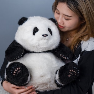 May include: A woman is holding a large, plush panda bear. The panda is white with black markings and has a black nose and eyes. The panda is wearing a black collar.
