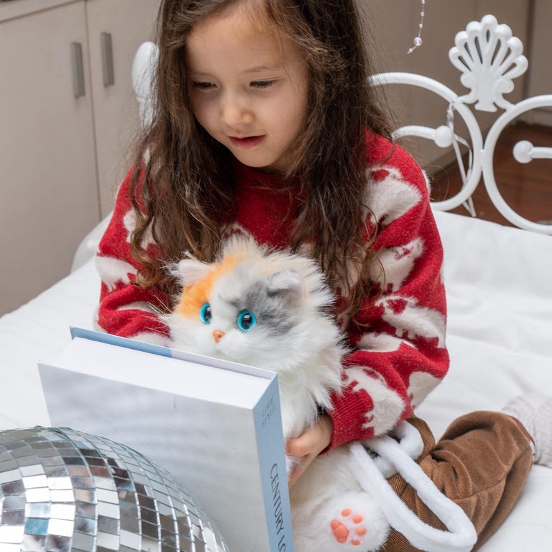 May include: A young girl in a red sweater with white sheep designs is sitting on a white bed with a white and gray plush cat. She is holding a book titled "Century of Love" in her lap. The cat has blue eyes and a pink nose.