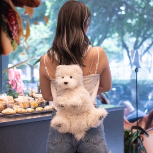 May include: A woman wearing a white tank top and blue denim shorts is carrying a white plush dog backpack. The dog is sitting upright with its paws resting on the woman's back.