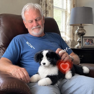 May include: A plush black and white dog toy with the text "wow~" and "woof~". The toy has a red heart graphic and is sitting on a person's lap. The person is wearing a blue shirt.