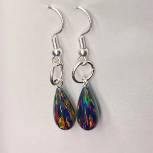 Könnte beinhalten: Ein Paar tropfenförmige Ohrringe mit schillernden, mehrfarbigen Mustern. Die Ohrringe haben silberfarbene Haken und Verbindungsringe. Die Tropfenanhänger zeigen eine dunkle Grundfarbe mit Streifen in Rot, Orange, Gelb und Grün.
