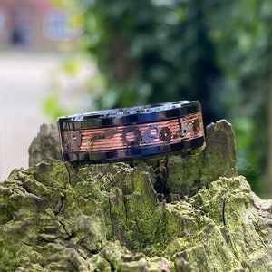 Puede incluir: Un anillo negro y cobre con un diseño detallado de engranajes y otros elementos mecánicos. El anillo está sobre un trozo de corteza de árbol.