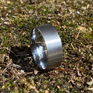 May include: A silver tungsten carbide ring with a brushed finish. The ring has the words "Tungsten Carbide" engraved on the side.