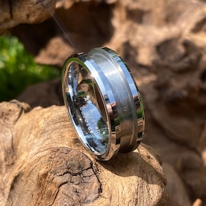 May include: A silver tungsten carbide ring with a brushed finish and a bevelled edge. The ring has a black inlay on the top and bottom of the band.