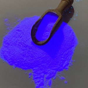 May include: A wooden scoop filled with bright blue glow-in-the-dark powder. The powder is scattered on a white surface.