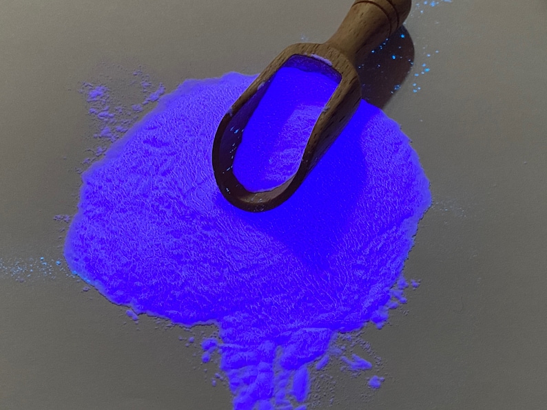 May include: A wooden scoop filled with bright blue glow-in-the-dark powder. The powder is scattered on a white surface.
