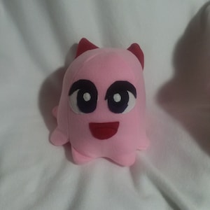 May include: Pink plush toy with a smiling face, large black eyes, and red horns. The toy is shaped like a ghost.