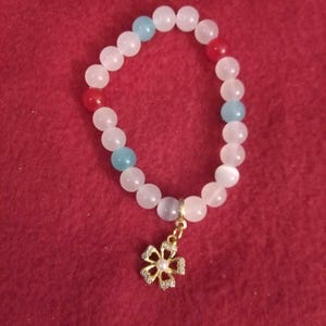 Puede incluir: Una pulsera de cuentas blancas, rojas y azules. Tiene un dije de flor dorada con una perla central y cristales transparentes. La pulsera se muestra sobre una superficie roja.