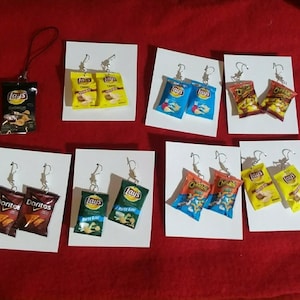 May include: A selection of novelty earrings, each designed as a small replica of a packet of crisps or snacks. Brands include Lay's, Doritos, and Cheetos. The earrings are attached to silver hooks and presented on white cards against a red backdrop.