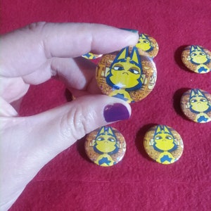 May include: Five round yellow and blue buttons with a cartoon cat wearing a gold crown and a pharaoh's headdress. The cat has a sad expression. The buttons are on a red surface.