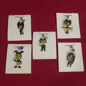 May include: Five different cartoon character charms on white card stock. The characters are all yellow with black outlines and are from the video game Bendy and the Ink Machine. The charms are attached to silver chains with bells.