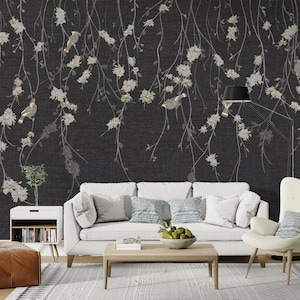 May include: A white sofa with blue and white patterned pillows sits in front of a black and white floral wallpaper with birds. The wallpaper features a repeating pattern of delicate branches with white flowers and small birds.