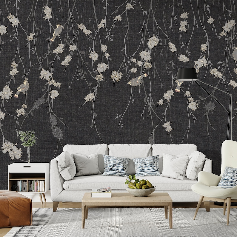 May include: A white sofa with blue and white patterned pillows sits in front of a black and white floral wallpaper with birds. The wallpaper features a repeating pattern of delicate branches with white flowers and small birds.