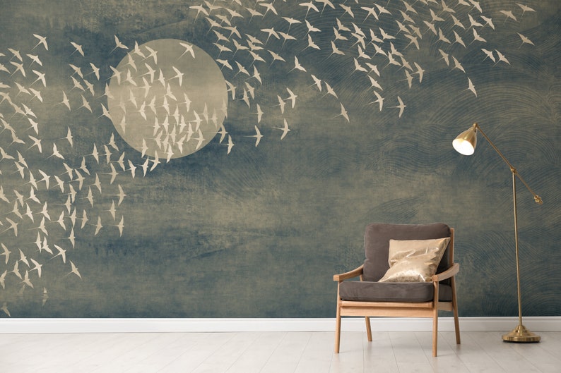 May include: A mural featuring a large, pale moon and a flock of birds in flight against a textured, dark blue background. A gray armchair with a gold pillow and a brass floor lamp complete the scene.