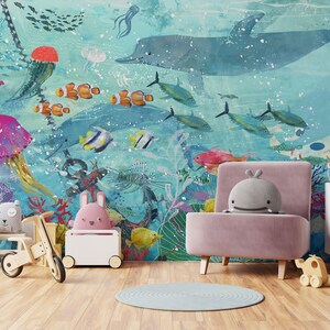 May include: A pink armchair with a gray whale plush toy sitting on it. The armchair is in front of a wall mural depicting an underwater scene with a blue whale, colorful fish, and coral reefs.