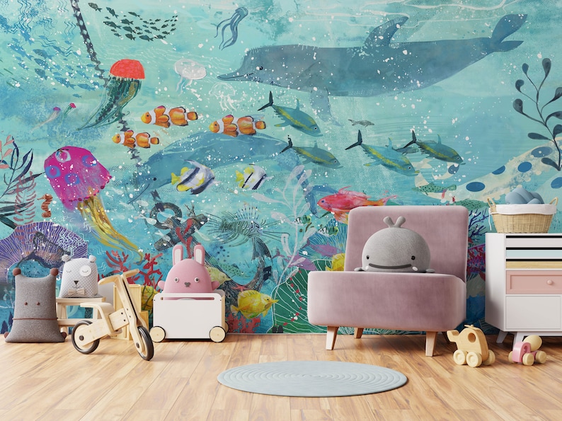 May include: A pink armchair with a gray whale plush toy sitting on it. The armchair is in front of a wall mural depicting an underwater scene with a blue whale, colorful fish, and coral reefs.