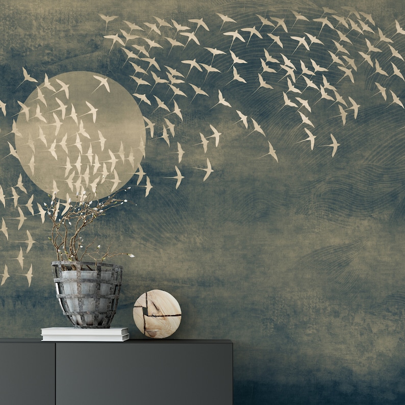 May include: A decorative wall mural featuring a stylized depiction of a full moon and a flock of birds in flight. The background has a textured, distressed appearance with shades of blue and beige. A decorative plant in a metal basket and a wooden disc are on a dark gray cabinet.