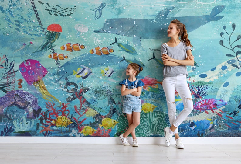May include: Two people stand in front of a blue and green underwater mural. The mural features a large dolphin, jellyfish, coral, and fish. The person on the left is a young girl wearing a blue denim overall dress. The person on the right is an adult woman wearing a grey shirt and white jeans.