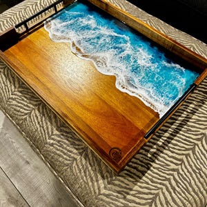 May include: A rectangular wooden serving tray with a beach-themed design. The tray features a wooden surface with a resin ocean scene in shades of blue and white. Black metal handles are attached to the sides. The tray is placed on a patterned ottoman.