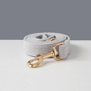 May include: A rolled-up light gray dog leash with a gold-colored metal clasp. The leash is made of a soft, textured material and is designed for pet owners. The background is a simple white and gray.