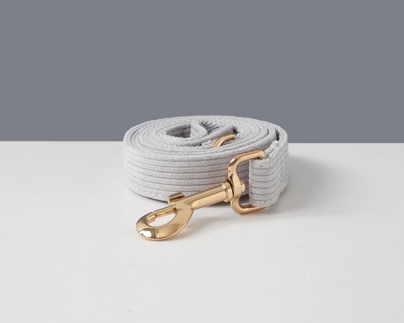 May include: A rolled-up light gray dog leash with a gold-colored metal clasp. The leash is made of a soft, textured material and is designed for pet owners. The background is a simple white and gray.