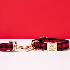 May include: Red and black buffalo plaid dog collar with a gold buckle and a personalized tag that reads "JM 02/12/2023".
